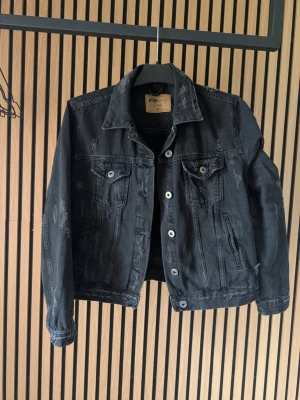 Svart jeansjacka med slitningar - Snygg svart jeansjacka med coola slitningar och silverfärgade knappar. Klassisk modell med två bröstfickor med lock och knapp. Jackan har en rak passform och är tillverkad i jeansmaterial. Perfekt för dig som gillar en edgy stil.