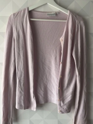 Ljuslila kofta från MAKEL - Söt ljuslila kofta från MAKEL med långa ärmar och rund hals. Koftan har en enkel design med små knappar framtill och är tillverkad i ett mjukt, tunt material som känns skönt mot huden. Perfekt att slänga över en topp för en chill look.