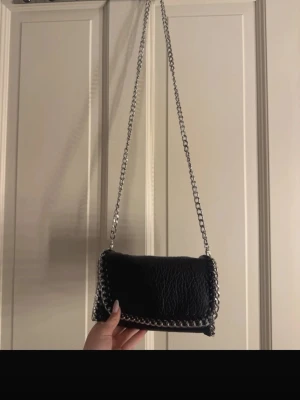 Svart axelväska med kedjedetaljer - Snygg svart axelväska i fejkläder med grov silverfärgad kedja som axelrem och runt kanterna. Väskan har en mjuk, lite puffig form och cool textur på materialet. Perfekt för dig som gillar en edgy och trendig look. använt en gång på bal, två fickor en dragkedja 
