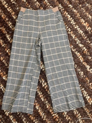 Rutiga vida byxor från KappAhl - Snygga vida byxor från KappAhl i rutigt mönster med svart, beige och vitt. Byxorna har hög midja med beige resår och är tillverkade i ett mjukt tyg som känns skönt mot huden. Perfekta för en trendig och avslappnad look.