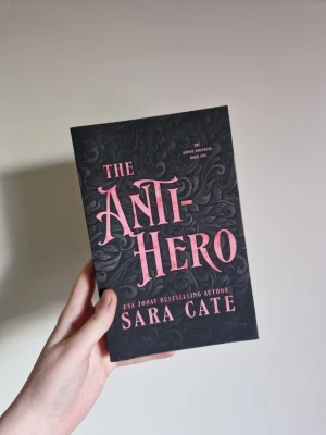The Anti-Hero av Sara Cate - En snygg skönlitterär bok i romantik genren med svart omslag och rosa text, skriven av Sara Cate. Boken har ett stilrent, mönstrat omslag och är första delen i The Good Brothers-serien. Helt ny och oläst. Katt finns i hemmet. Nypris 204 kronor