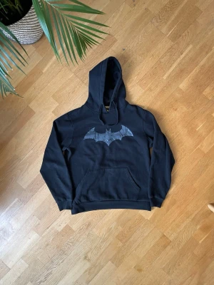 Svart Batman hoodie med tryck - Svart hoodie med stort Batman-logga tryckt på bröstet. Klassisk känguruficka framtill och huva med dragsko. Mjuk bomullskänsla och ribbade muddar vid ärmslut och nederkant. Perfekt för dig som gillar superhjältar och streetwear. Väldigt skön att gymma i. Storlek L men sitter som M.
