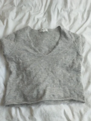 Grå stickad topp från Gina Tricot - Mjuk och fluffig grå stickad topp från Gina Tricot i alpackablandning. Modellen har korta ärmar och djup v-ringning, perfekt att styla med jeans eller kjol. Enkel och stilren design som passar till många outfits.