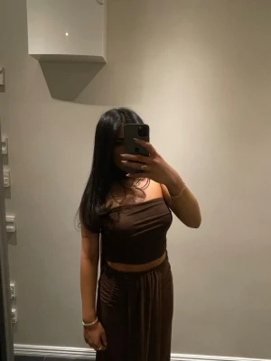 Brunt tvådelat set med scarf - Säljer ett brunt tvådelat set med en axelbandslös crop top och matchande höga byxor med dragsko i midjan. Kommer med en scarf som in visas på bilderna. Setet har en tight passform och är gjort i ett mjukt, stretchigt tyg som sitter snyggt på kroppen. Perfekt för dig som gillar stilrena och trendiga plagg.