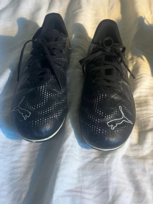 Svarta Puma fotbollsskor med dobbar - Säljer ett par svarta fotbollsskor från Puma med vita detaljer och logga på tån. Skorna har snörning och dobbar för bra grepp på planen. Ovandelen är i syntetmaterial med mönstrade vita prickar för extra stilpoäng. Perfekta för dig som vill sticka ut på matchen!