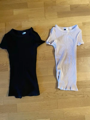 2 ribbade toppar från Divided H&M - Två ribbade toppar från Divided H&M i storlek XS. En är svart och den andra är rosa. Båda har korta ärmar och rund halsringning, perfekta att styla med jeans eller kjol. Materialet är mjukt och stretchigt för en bekväm passform.