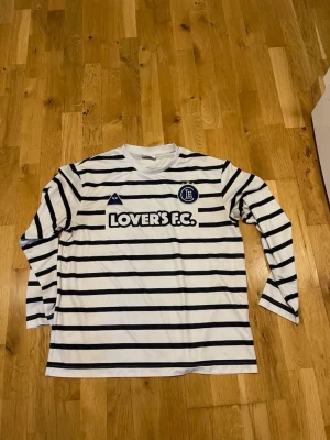HM Randig långärmad tröja Lover's F.C. Stl M - Snygg långärmad tröja från Lover's F.C. i vitt med svarta horisontella ränder. Tryckt logga och text på bröstet samt nummer 12 på ryggen. Tillverkad i mjukt och bekvämt material, perfekt för sport eller streetwear.