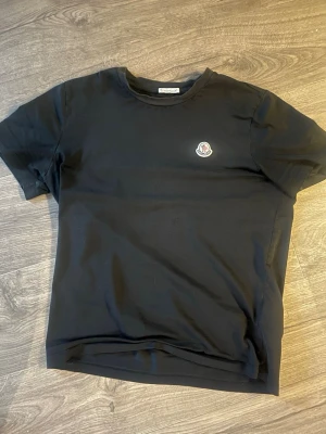 Svart Moncler t-shirt med logga - Svart t-shirt från Moncler med klassisk rund hals och broderad logga på bröstet. Enkel och stilren design som passar perfekt till jeans eller shorts. Tillverkad i mjuk bomull för en bekväm känsla hela dagen.
