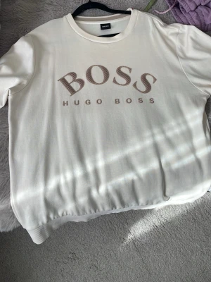 Vit sweatshirt från Hugo Boss - Säljer en stilren vit sweatshirt från Hugo Boss med stor BOSS-logga och texten HUGO BOSS framtill. Tröjan har rund halsringning, ribbade muddar och är gjord i mjukt material. Perfekt för en clean och trendig look.