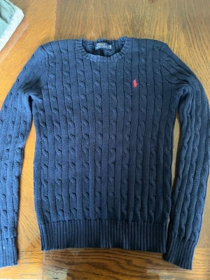 Mörkblå kabelstickad tröja Polo Ralph Lauren - Mörkblå kabelstickad tröja från Polo Ralph Lauren med rund halsringning och klassisk röd broderad logga på bröstet. Tröjan har långa ärmar och ribbade muddar vid ärmslut och nederkant. Perfekt för en stilren och tidlös look.