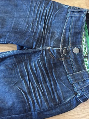 Blå jeans från Sweet Years - Säljer ett par blå jeans från Sweet Years med slitna detaljer och markerade veck framtill. Jeansen har dubbla knappar i midjan, grönt broderi på bakfickan och klassisk femficksmodell. Snygg kontrastsöm och cool tvättad look.