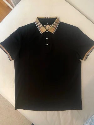Burberry pike - Svart pikétröja från Burberry med klassisk rutig krage och matchande detaljer vid ärmsluten. Tröjan har tre knappar framtill och är tillverkad i mjuk bomull. Perfekt för en stilren och trendig look.