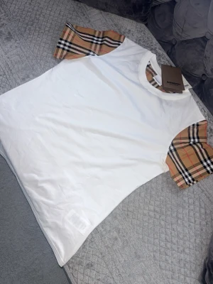 Vit Burberry t-shirt med rutiga ärmar - Snygg vit t-shirt från Burberry med klassiska beige-rutiga ärmar och halsringning. Perfekt för dig som vill ha en stilren look med en touch av lyx. Tillverkad i mjuk bomull och har en normal passform.