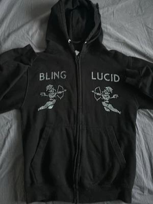 Svart hoodie från Bling Lucid med tryck - Svart zip-hoodie från Bling Lucid med huva och dragsko. På bröstet finns vita tryck med texten 'BLING LUCID' och två cupido-figurer. Hoodien har fickor framtill och är tillverkad i mjukt material, perfekt för en chill och stilren look.