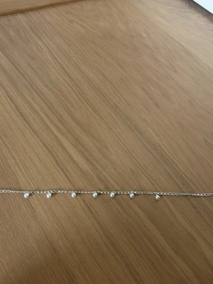 Silverfärgat halsband med pärlor - Snyggt halsband i silverfärg med små vita pärlor som hänger längs kedjan. Kedjan är tunn och har ett enkelt, stilrent uttryck. Perfekt för dig som gillar minimalistiska smycken med en touch av elegans.