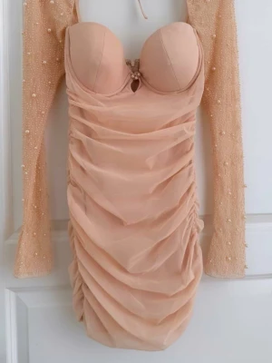 Beige kort klänning med pärldetaljer - Supersnygg beige kort klänning med vadderad byst, rynkade detaljer och långa ärmar täckta av pärlor. Klänningen har en figurnära passform och meshmaterial som ger en lyxig känsla. Perfekt för dig som vill sticka ut med stilrena detaljer. Byst är ungefär en D kupa