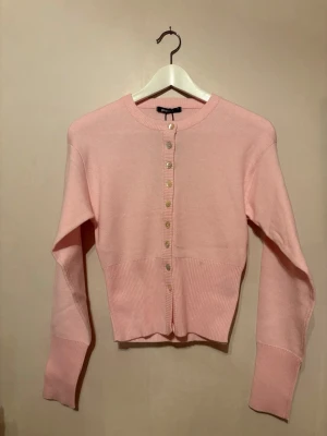 Ljusrosa kofta med knappar - Söt ljusrosa kofta med rund hals och knappar framtill. Koftan har långa ärmar och bred ribbad mudd vid midjan och ärmsluten. Perfekt för att addera en mjuk touch till din outfit.