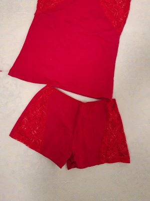 Röd pyjamas med spetsdetaljer - Snygg röd pyjamas i två delar med linne och shorts. Båda delarna har fina spetsdetaljer på sidorna som ger en extra touch. Materialet ser mjukt och stretchigt ut, perfekt för att chilla hemma. Inget synligt märke på plagget.