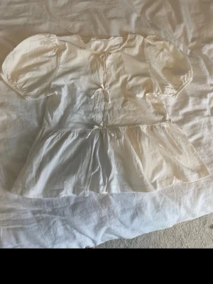 Vit blus med puffärmar och knyt - Söt vit blus med puffiga korta ärmar och två dekorativa knyt framtill. Blusen har en lös passform med volang nedtill som ger en romantisk vibe. Tillverkad i ett tunt, mjukt bomullstyg som känns luftigt och skönt. I storlek 170 men sitter som XS. Använd få gånger