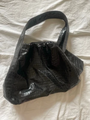 Svart axelväska i krokopräglat material - Säljer en svart handväska med bred axelrem och krokopräglat mönster. Väskan har en mjuk, slouchy form och dragskostängning upptill. Perfekt för dig som gillar trendiga accessoarer med lite edge.
