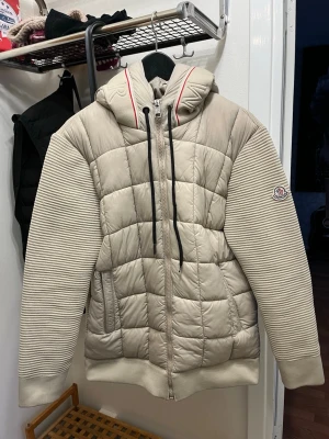 Beige dunjacka med huva från Moncler - Snygg beige dunjacka från Moncler med quiltad kropp och stickade ärmar. Jackan har huva med dragsko, hel dragkedja framtill och Moncler-logga på ärmen. Perfekt för kalla dagar och riktigt trendig look.