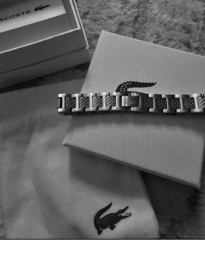 Silverfärgat armband från Lacoste - Snyggt armband i silverfärgad metall från Lacoste med rektangulära länkar och graverade detaljer. Armbandet har en stilren design med Lacostes ikoniska krokodil på låset. Perfekt accessoar för dig som gillar minimalistisk och modern stil.