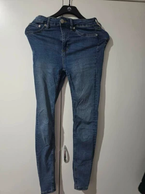 Blå skinny jeans med hög midja - Säljer ett par klassiska blå skinny jeans med hög midja och fem fickor. Jeansen har dragkedja och knapp framtill samt bälteshällor. Materialet är stretchigt denim som sitter tight och bekvämt. Perfekta till en avslappnad streetstyle-look.