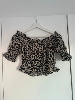 Leopardmönstrad offshoulder topp - Säljer en leopardmönstrad offshoulder topp med volangdetaljer och knappar framtill. Toppen har korta ärmar med resår och är croppad i modellen. Perfekt för dig som vill sticka ut lite extra.