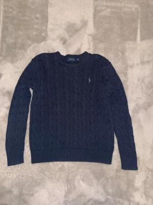 Mörkblå kabelstickad tröja Polo Ralph Lauren - Kabelstickad tröja i mörkblått från Polo Ralph Lauren med rund halsringning och diskret broderad logga på bröstet. Tröjan har ribbade muddar vid ärmslut och nederkant, perfekt för dig som gillar klassisk och stilren look.