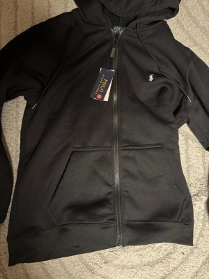 Svart hoodie från Polo Ralph Lauren - Svart hoodie med dragkedja från Polo Ralph Lauren. Klassisk design med huva, två fickor framtill och den ikoniska lilla logotypen broderad på bröstet. Tillverkad i mjukt bomullsmaterial som känns skönt mot huden. Perfekt för en avslappnad och stilren look. HELT NY