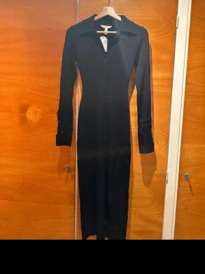 Svart långärmad klänning med knappar - Stilren svart långklänning med lång ärm och knappar hela vägen framtill. Klänningen har en klassisk krage och figurnära passform som ger en snygg siluett. Perfekt för dig som gillar minimalistisk och tidlös stil.