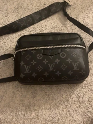 Svart axelväska  - Snygg svart axelväska från med klassiskt monogrammet i grått. Väskan har justerbar axelrem, dubbla dragkedjor och är tillverkad i skinn. Perfekt för dig som vill ha en stilren och lyxig accessoar. Svåra att hitta för låga priser. 