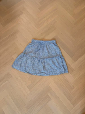 Blå blommig volangkjol - Söt blå kortkjol med små vita blommor och volangdetalj. Kjolen har resår i midjan för en bekväm passform och är perfekt för varma dagar. Lätt och luftig modell som ger en avslappnad och trendig look. Storlek Xs ungefär men passar som S