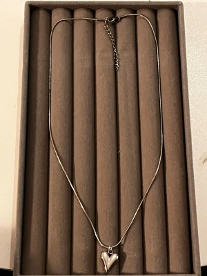 Silverhalsband med hjärthänge - Snyggt halsband i silverfärgad metall med ett blankt hjärthänge. Kedjan är tunn och har ett justerbart lås med förlängningskedja. Perfekt accessoar för att lyfta vilken outfit som helst. Hjärtat ger en romantisk touch och halsbandet är enkelt att matcha.