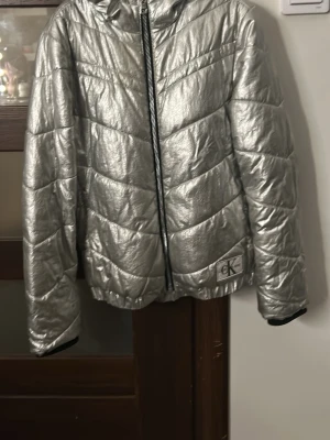Silver pufferjacka från Calvin Klein - Snygg och glansig silverfärgad pufferjacka från Calvin Klein Jeans. Jackan har huva, dragkedja framtill och ett stort CK-märke vid nederkanten. Quiltad design med breda sömmar och mjukt, vadderat material som håller dig varm under vintern.
