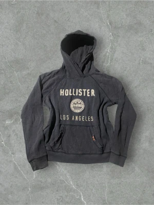 Mörkblå hoodie från Hollister LA | 40 - Mörkgrå hoodie från Hollister med stor huva och magficka. Framtill står det 'HOLLISTER LOS ANGELES' i beige bokstäver och ett tryck med palmer och texten 'California Since 1922'. Skön collegekänsla och ribbade muddar vid ärmar och nederkant.