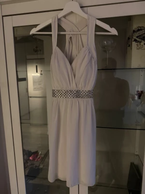 Vit kortklänning med paljettbälte - Supersnygg vit kortklänning med djup v-ringning och brett paljettbälte i midjan. Klänningen har ett elegant axelbandsdesign med kors i ryggen och ett mjukt, flytande tyg som ger en lyxig känsla. Perfekt för dig som vill sticka ut!