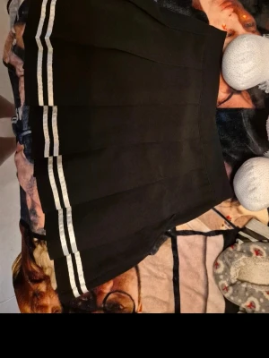 Svart plisserad kjol  - Snygg svart plisserad kjol från SHEIN med vita ränder längs nederkanten. Kjolen har en kort modell och är perfekt för en trendig och sportig look. Tillverkad i ett mjukt syntetmaterial som ger bra passform och komfort.