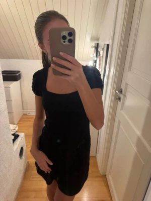 Svart virkad klänning med puffärm - Snygg svart klänning med virkat mönster och fyrkantig urringning. Klänningen har korta puffärmar och sitter figurnära upptill. 