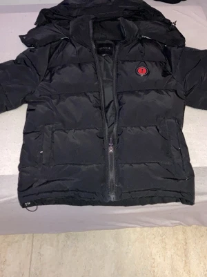 Trapstar jacka  - Säljer en svart pufferjacka från Topstreetwear med huva och dragkedja framtill. Jackan har två stora fickor och en röd patch med texten 'It's a Secret' på bröstet. Perfekt för kalla vinterdagar och riktigt skön att bära.