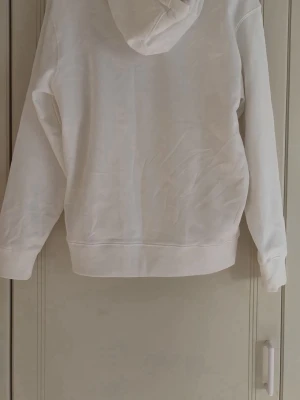 Vit Dsquared2 ICON hoodie - Säljer en vit hoodie från Dsquared2 med stor svart 'ICON'-text och färgstänk i grönt, rött och blått på bröstet. Hoodien har huva och en stor magficka framtill. Perfekt för dig som gillar streetwear och vill sticka ut lite extra. Använd 1 gång då jag fortfarande har kvar tagen.