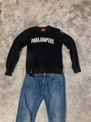 Svart Parajumpers sweatshirt - Svart sweatshirt från Parajumpers med vit logga tryckt över bröstet. Tröjan har rund halsringning, långa ärmar och ribbade muddar vid ärmslut och nederkant. Klassisk och stilren design med patch på ena ärmen.