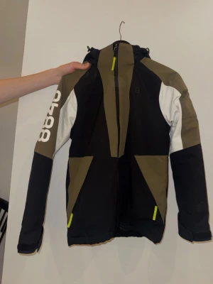 Svart och olivgrön jacka 8848 Altitude - Snygg vindjacka från 8848 Altitude i svart, olivgrönt och vitt . Jackan har huva, hel dragkedja framtill och två fickor med dragkedja. Stora vita siffror på ena ärmen och logga på bröstet. I väldigt bra skick! 