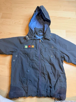 Mörkblå vindjacka med broderi - Mörkblå vindjacka med huva och dragkedja framtill. Jackan har broderade detaljer på ryggen med djur och spår, samt färgglada märken på bröstet. Praktiska fickor med knappar och ribbade muddar vid ärmslut och nederkant. Perfekt för kyliga dagar.