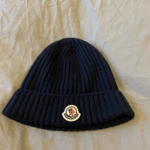 Mörkblå ribbstickad Moncler mössa - Snygg mörkblå ribbstickad mössa från Moncler med klassisk uppvikt kant och det ikoniska Moncler-logotypen framtill. Tillverkad i mjukt och varmt material som passar perfekt för kyliga dagar. Enkel men stilren design som funkar till det mesta.