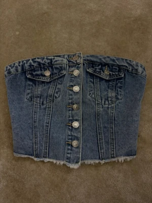 Denim topp - Snygg tubtopp i blå denim med knappar framtill och rå kant nertill. Toppen har två fejkfickor med lock och smock baktill för extra stretch. Perfekt för dig som vill ha en edgy och trendig look. Dee jeans matrial och är helt ny