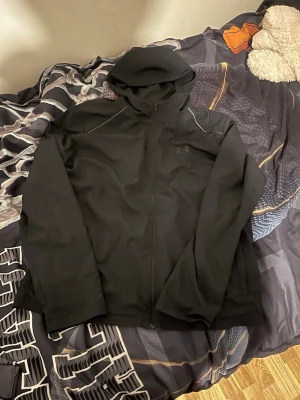 Svart hoodie från Under Armour - Snygg svart hoodie från Under Armour med dragkedja framtill och stor huva. Diskret logga på bröstet och stort UNDER ARMOUR-tryck på ryggen. Tillverkad i ett mjukt och stretchigt material som passar perfekt till chill eller träning.