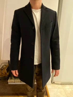 Svart rock från J.Lindeberg, strl 44 - Säljer en stilren svart rock från J.Lindeberg i storlek 44. Väldigt fint skick. Klassisk modell med tre knappar framtill, diskret bröstficka och slät finish. Yttertyget är i ullblandning och fodret i viskos, vilket gör den både varm och bekväm. Perfekt för höst och vinter.