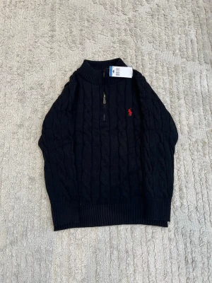 Svart kabelstickad tröja Polo Ralph Lauren - Kabelstickad svart tröja från Polo Ralph Lauren med halv dragkedja och röd broderad logga på bröstet. Tröjan har hög krage och ribbade muddar vid ärmslut och nederkant. Perfekt för en stilren och klassisk look.