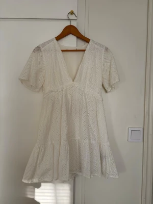 Vit studentklänning  med v-ringning - Superfin vit klänning med v-ringning och korta puffärmar. Klänningen har broderade spetsdetaljer i ett snyggt mönster och volang nertill. Perfekt för sommarens alla tillfällen och har en luftig passform.